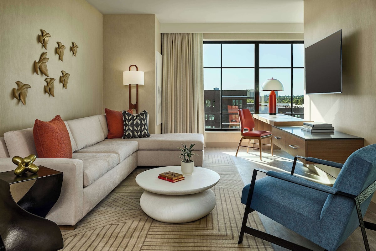 A modern hotel room with a sectional sofa, a round coffee table, a blue armchair, a desk with a chair, and a large window with a city view.
