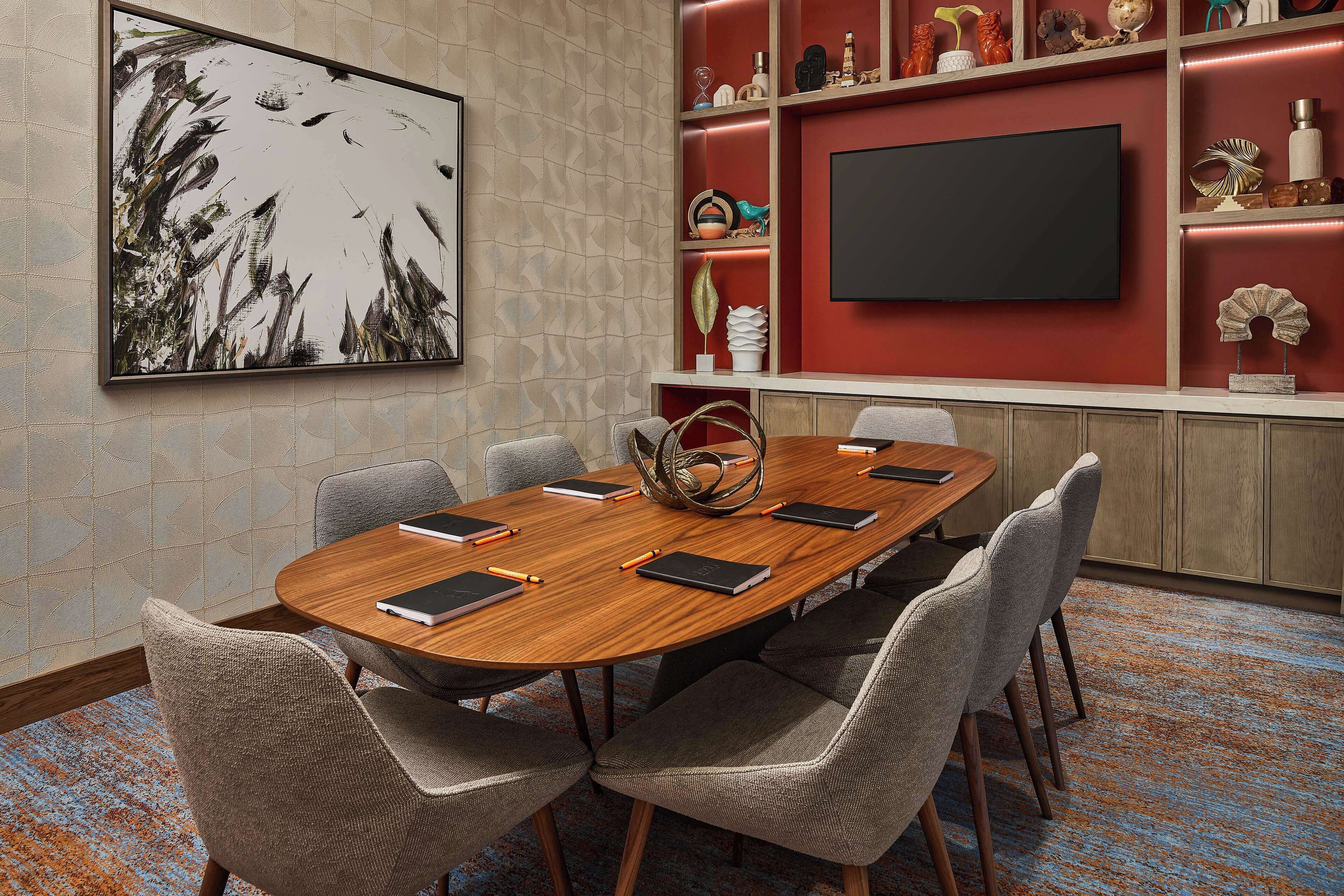 Un salón de conferencias con una mesa de madera, seis sillas, un cuadro grande, un televisor y estantes con objetos decorativos.