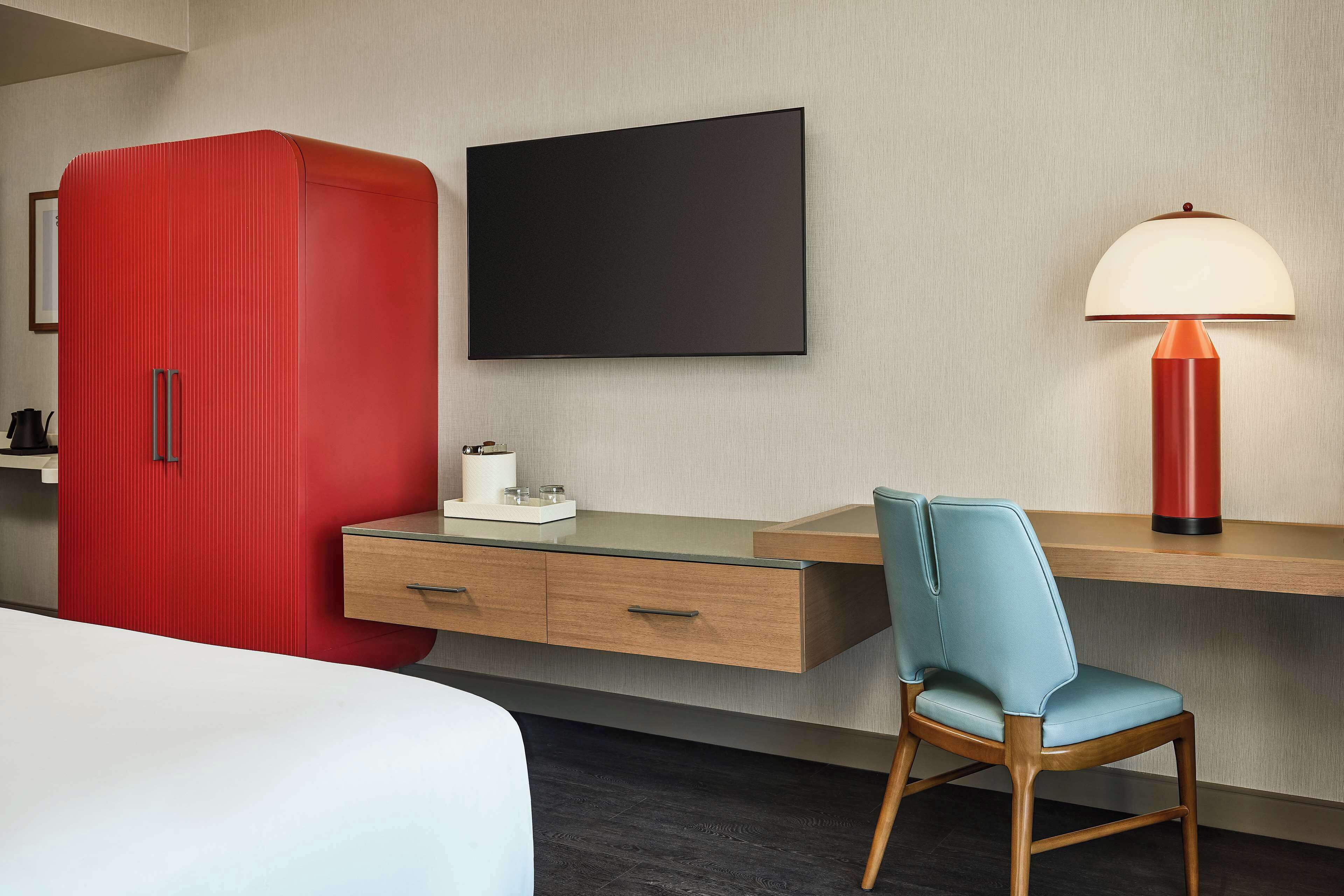 A hotel room with a bed, a desk with drawers, a red wardrobe, a flat-screen TV, a lamp, and a blue chair.