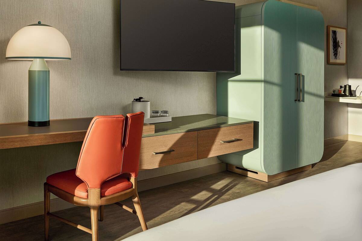 A modern hotel room with a wooden desk, a red chair, a flat-screen TV, a teal wardrobe, and a lamp.