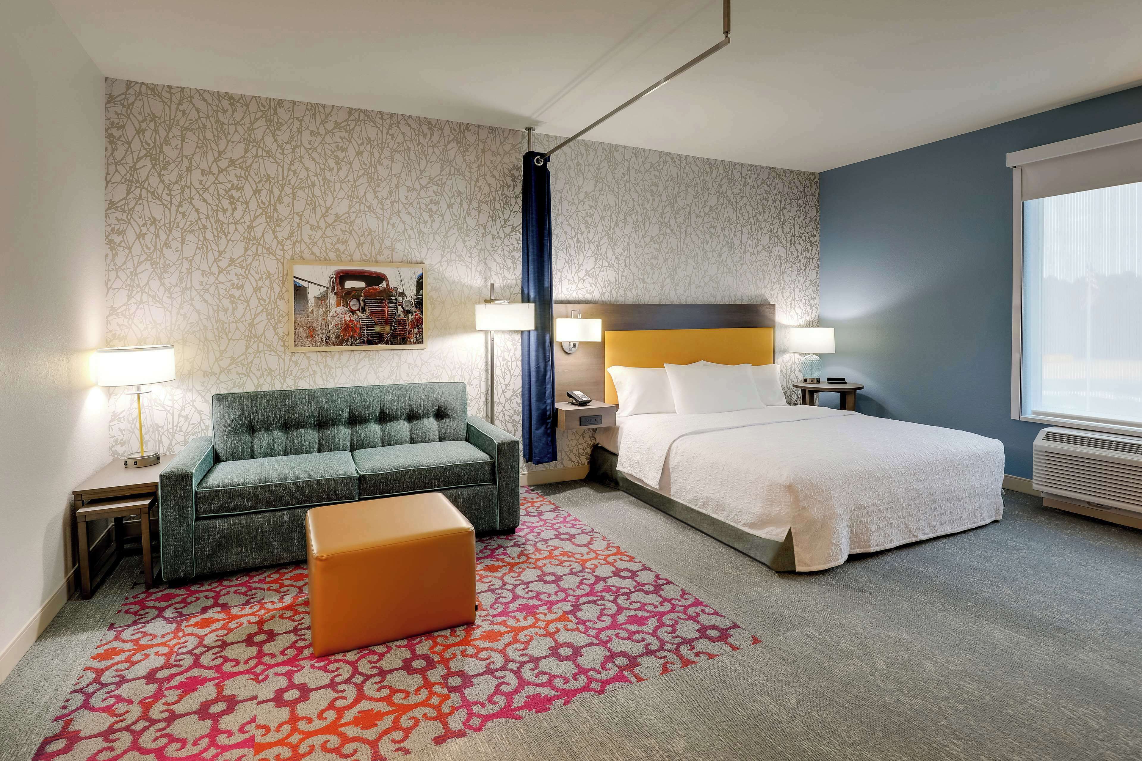 A hotel room with a bed, a sofa, a nightstand, a lamp, a patterned rug, and a window.