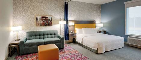 A hotel room with a bed, a sofa, a nightstand, a lamp, a patterned rug, and a window.