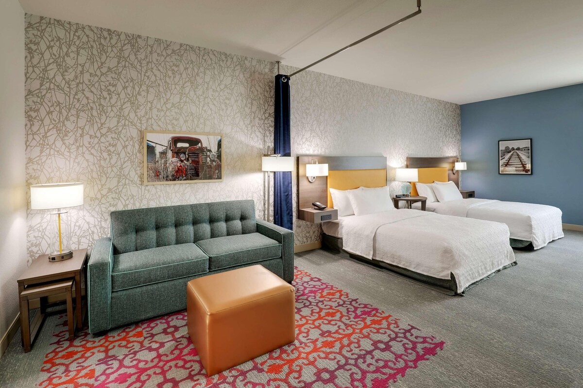 Una habitación de hotel con un sofá, dos camas, una alfombra estampada y una mesita de noche con lámpara.