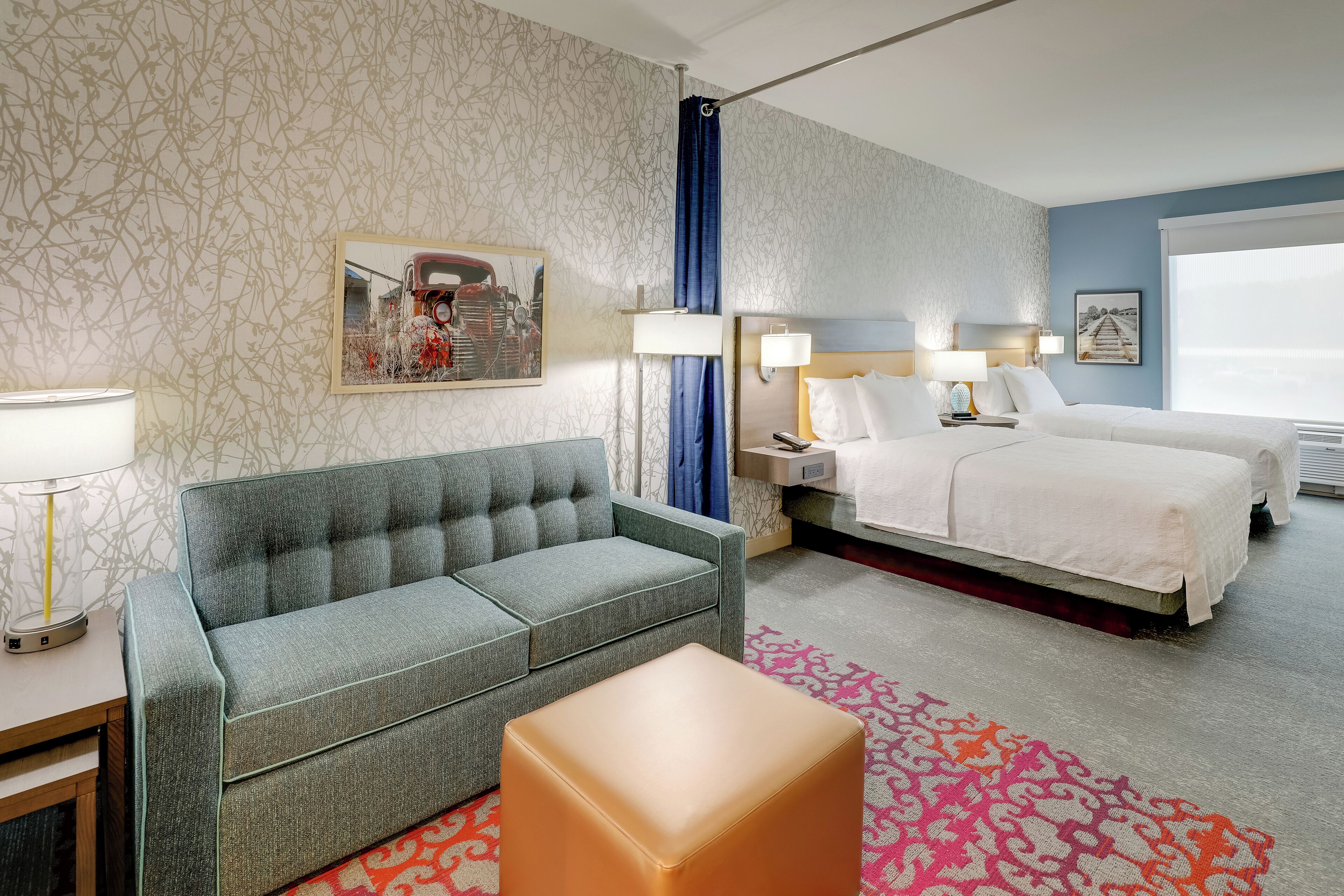 A hotel room with a grey sofa, a bed with white linens, a nightstand with lamps, and a large window.