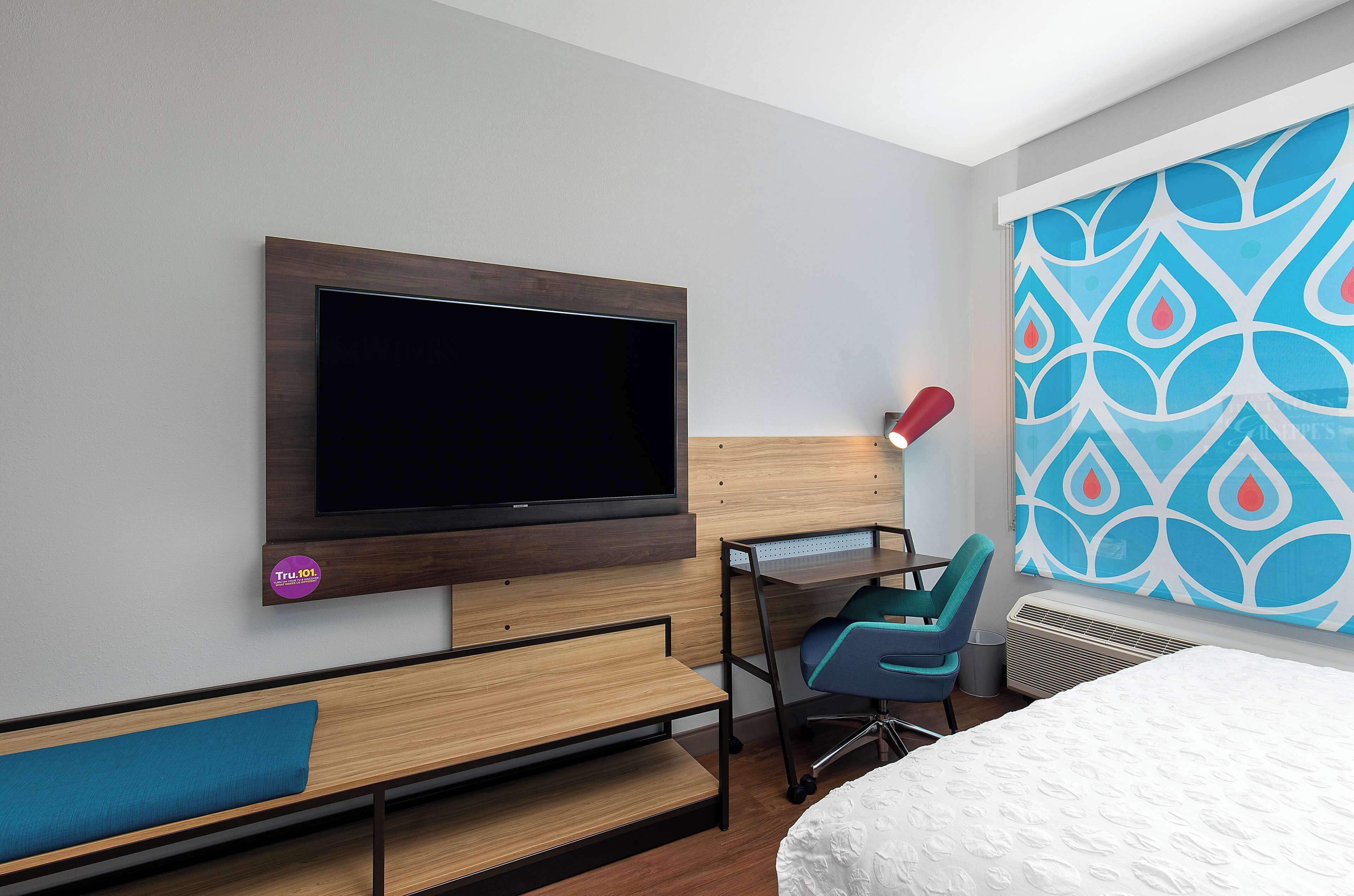A modern hotel room with a large flat-screen TV mounted on the wall, a desk with a chair, a bed with a white mattress, and a colorful abstract wall design.