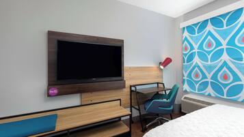 A modern hotel room with a large flat-screen TV mounted on the wall, a desk with a chair, a bed with a white mattress, and a colorful abstract wall design.