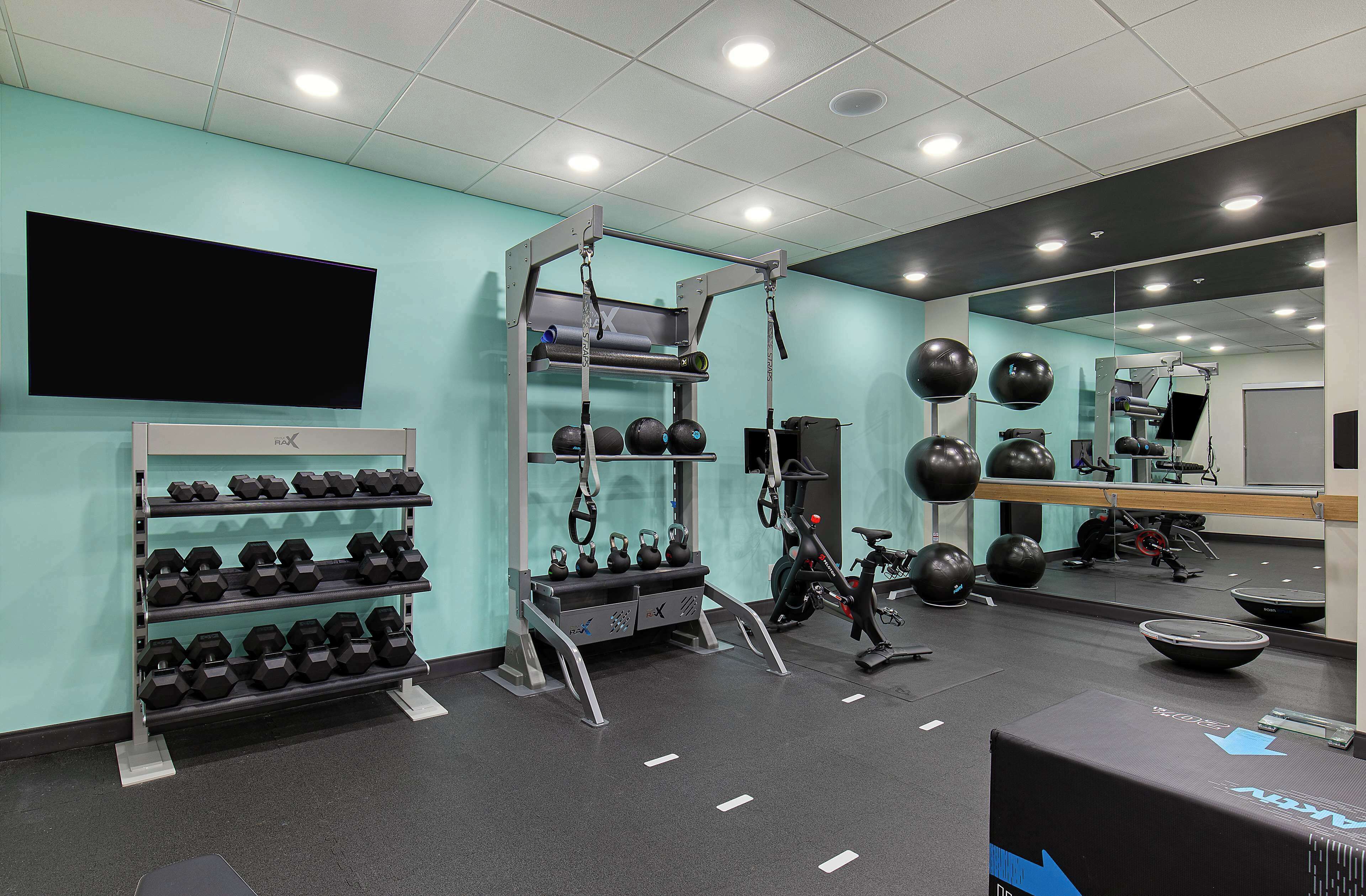 A gym with weightlifting equipment, a wall-mounted TV, and a mirror reflecting the space.