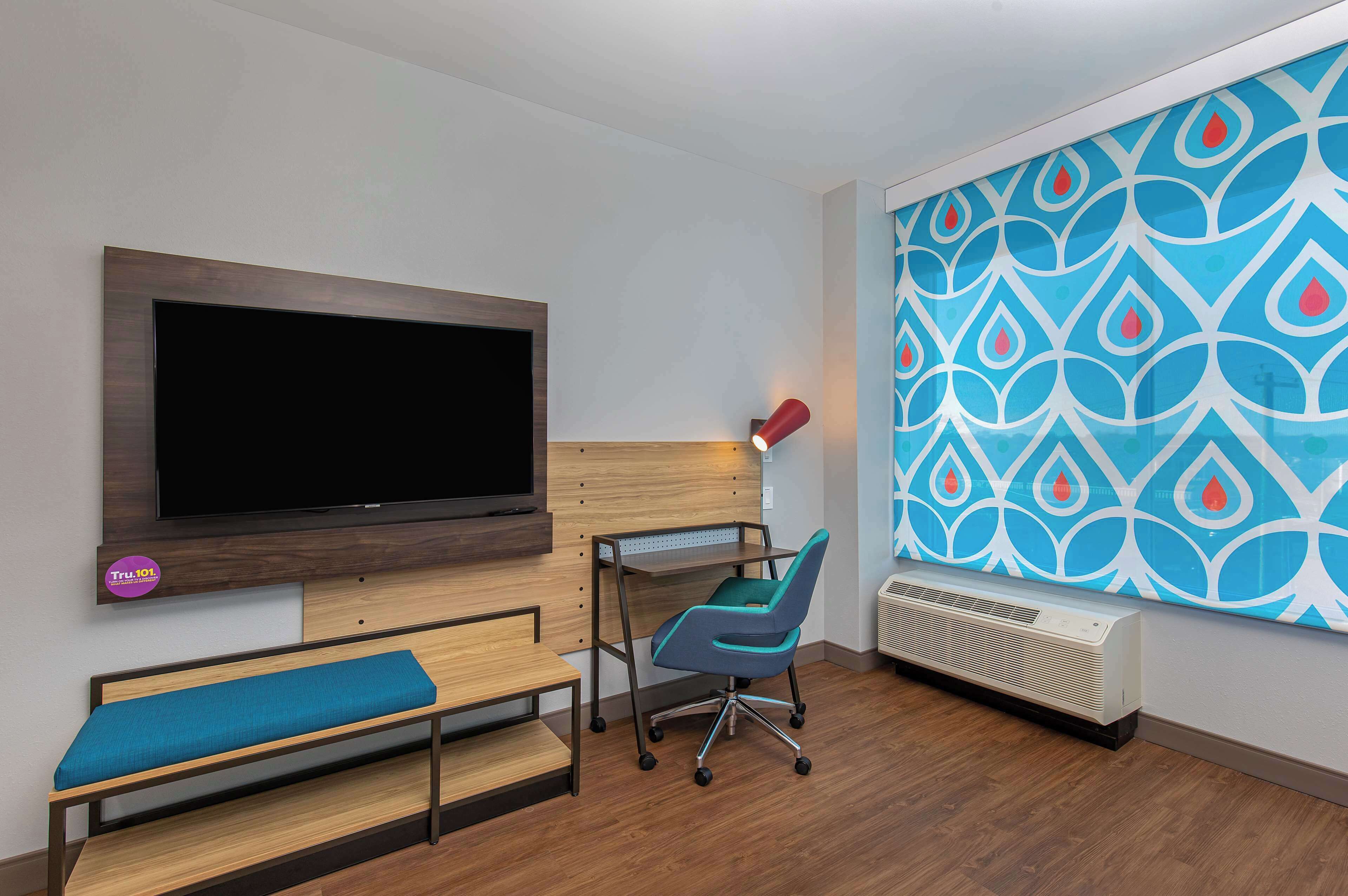 A modern room with a flat-screen TV, a desk with a chair, a bench, a radiator, and a decorative wall with a geometric pattern.