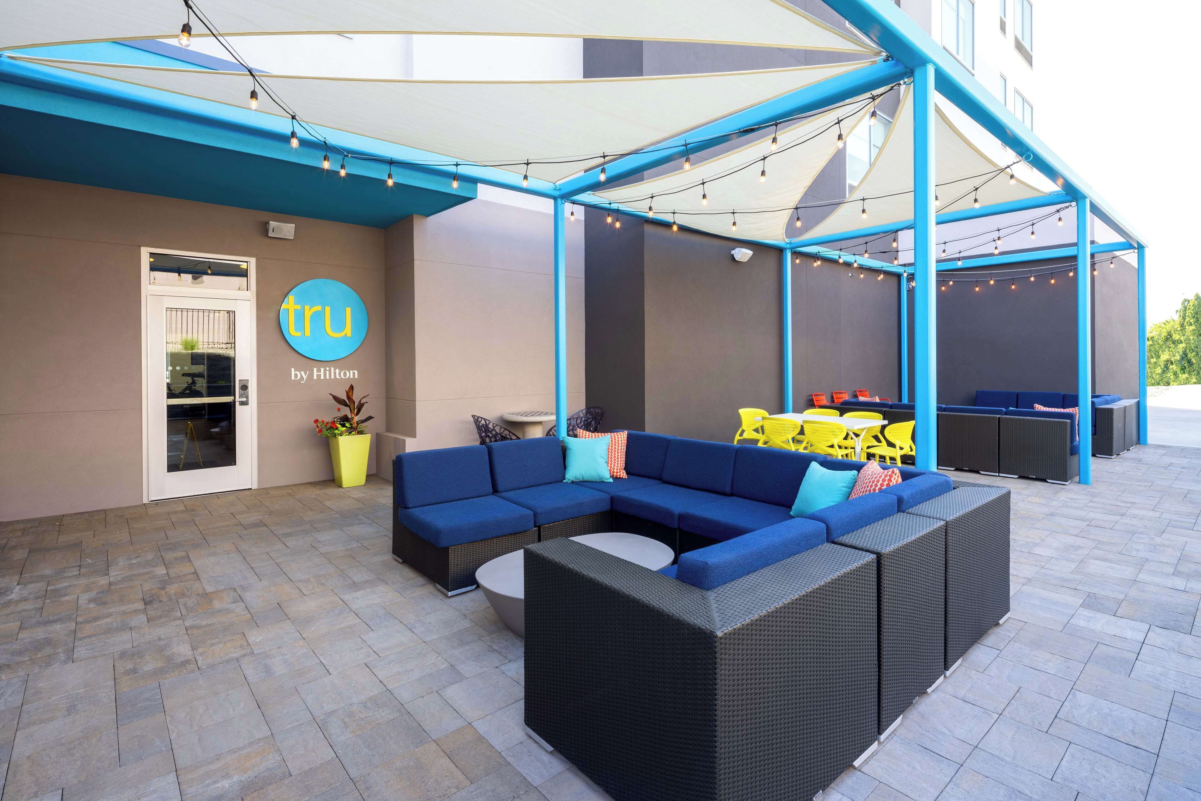 Un espace extérieur aménagé avec des meubles en osier bleu et noir, une table jaune et une porte blanche portant une enseigne indiquant « tru by Hilton ».