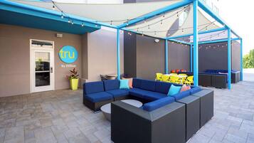 Un espace extérieur aménagé avec des meubles en osier bleu et noir, une table jaune et une porte blanche portant une enseigne indiquant « tru by Hilton ».