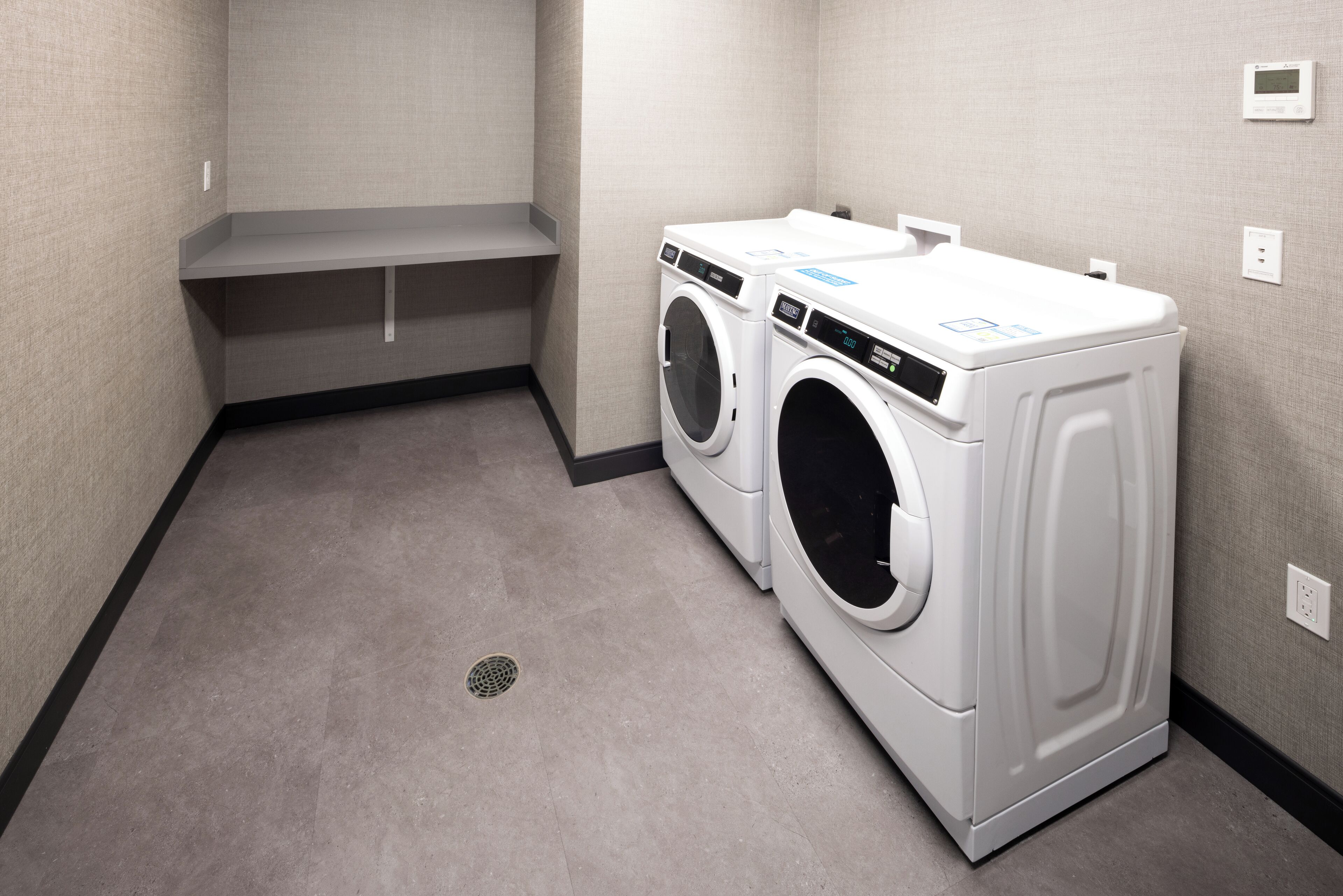 A laundry room with two white front-loading washing machines, a grey bench, and a vent on the wall.