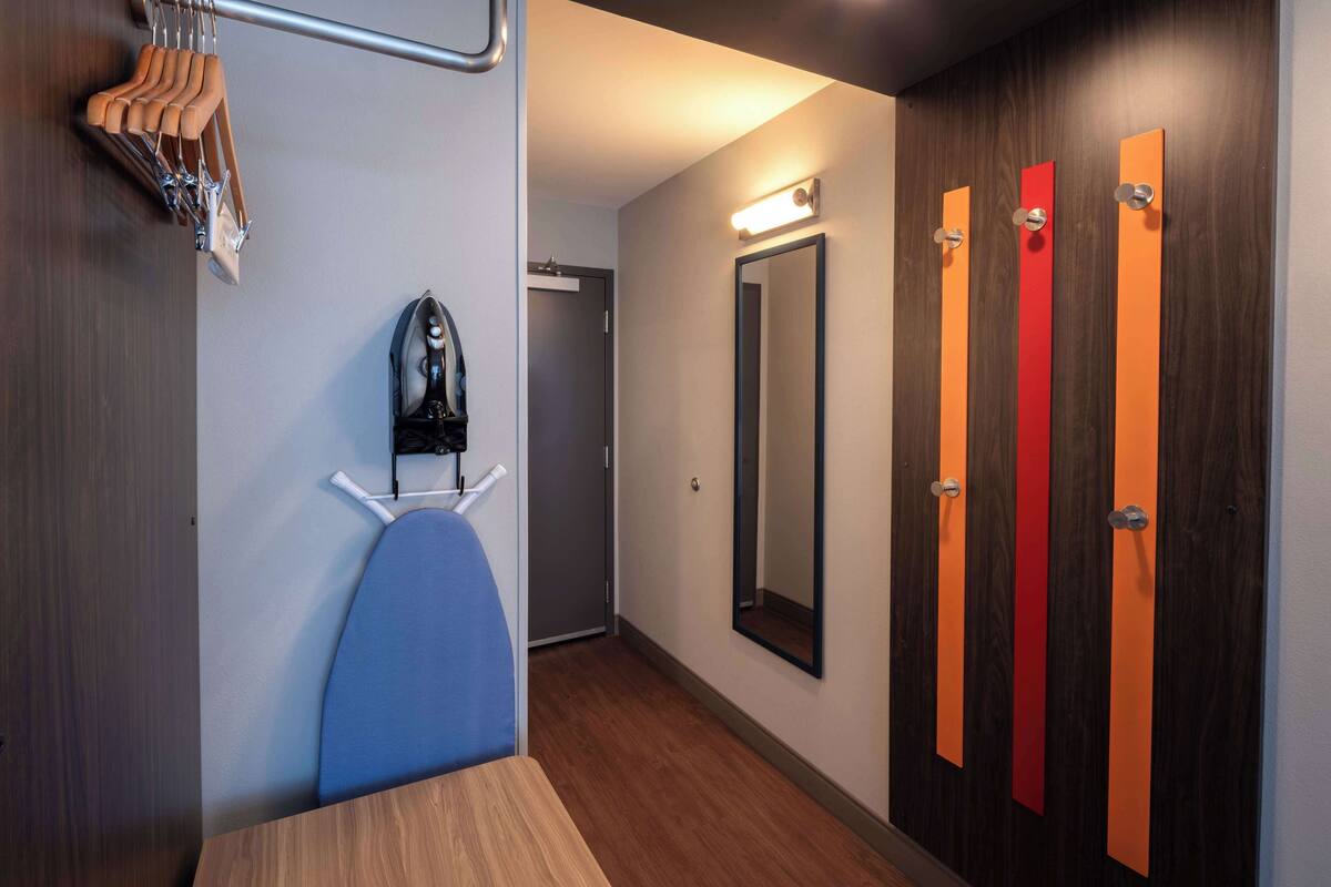 A hotel room with a wooden floor, a blue chair, a shoe rack, and a mirror.