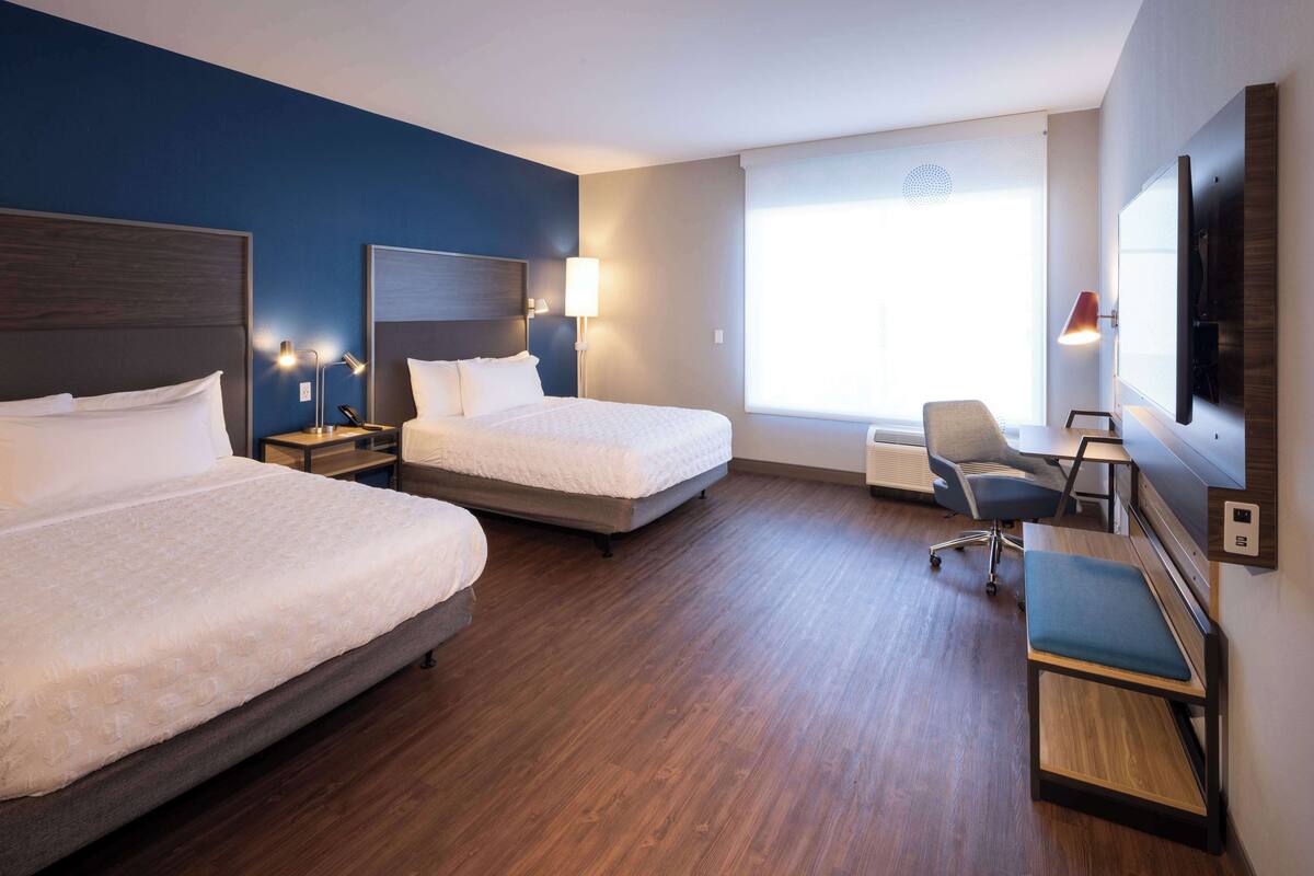 A hotel room with two beds, a desk, a chair, a TV, and a large window.