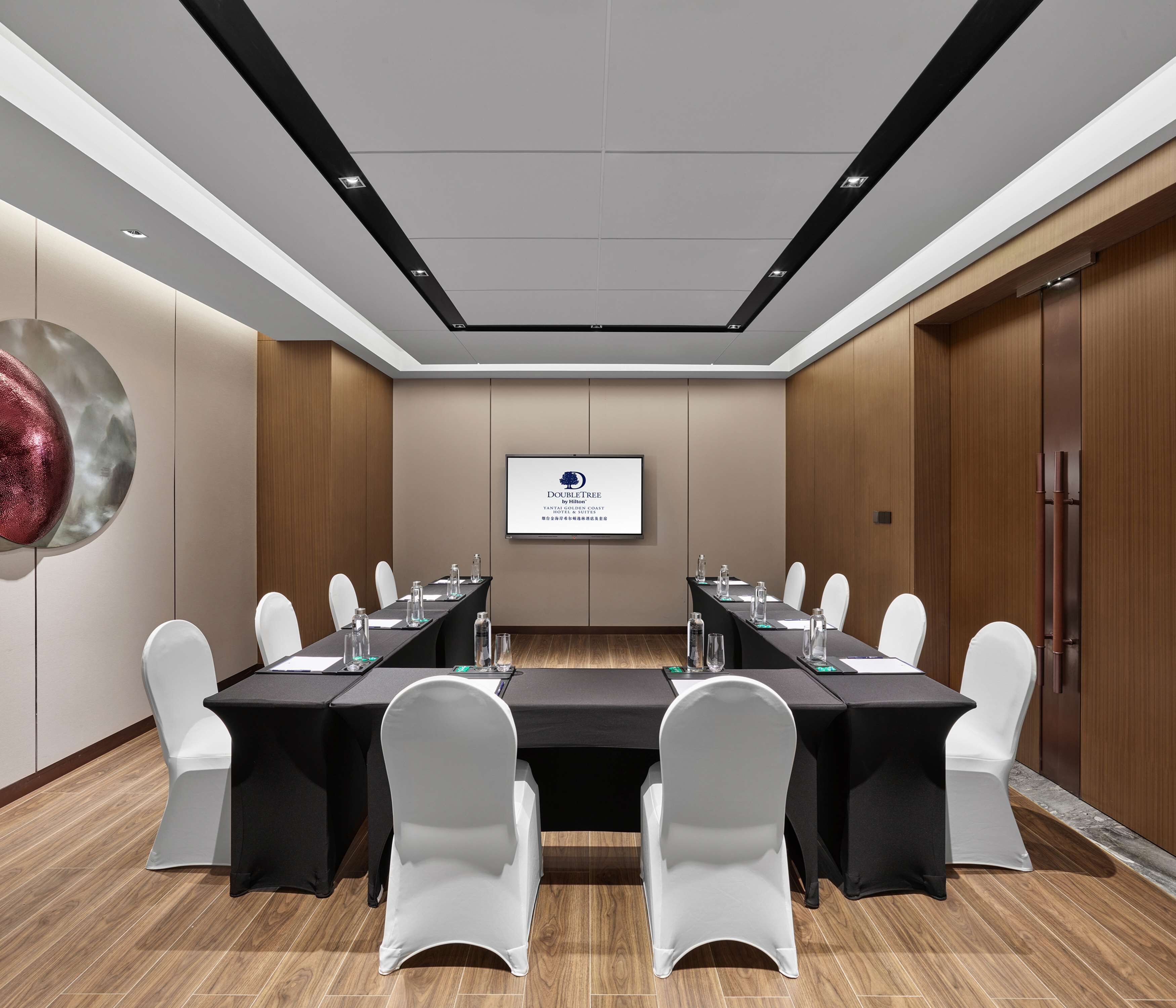 Un salón de conferencias con una larga mesa, sillas blancas y una pantalla que muestra un logotipo.