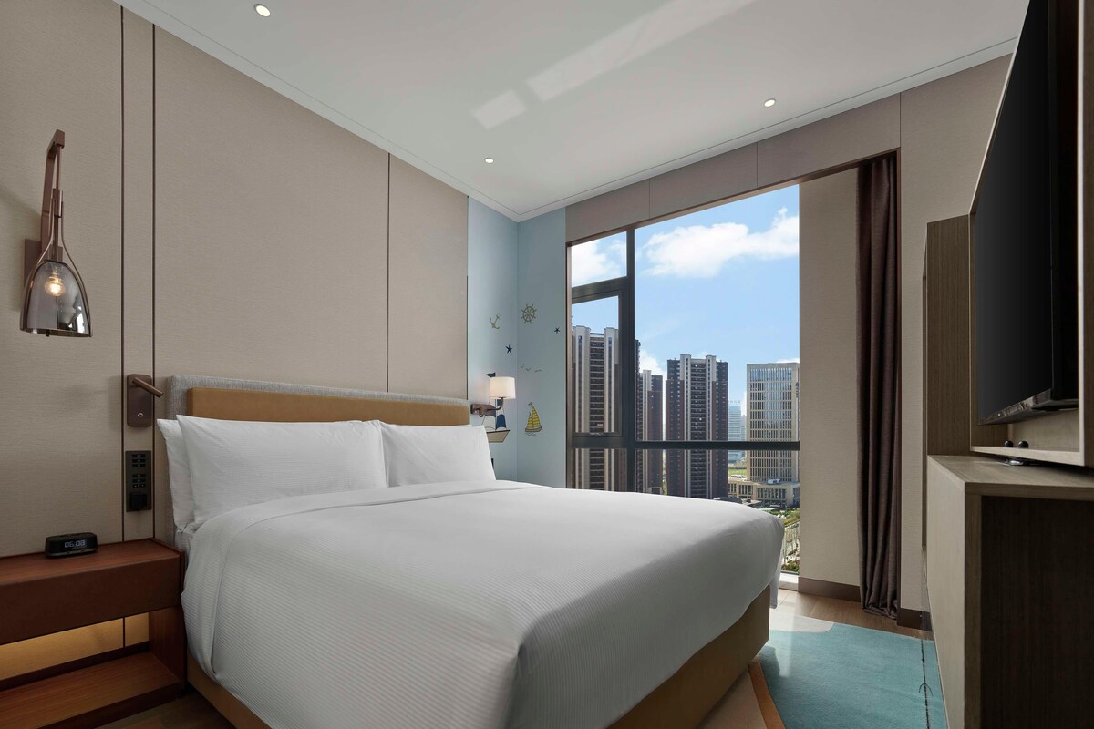 Una habitación de hotel moderna con una cama grande, una mesita de noche con lámpara, un televisor y vistas a edificios altos.