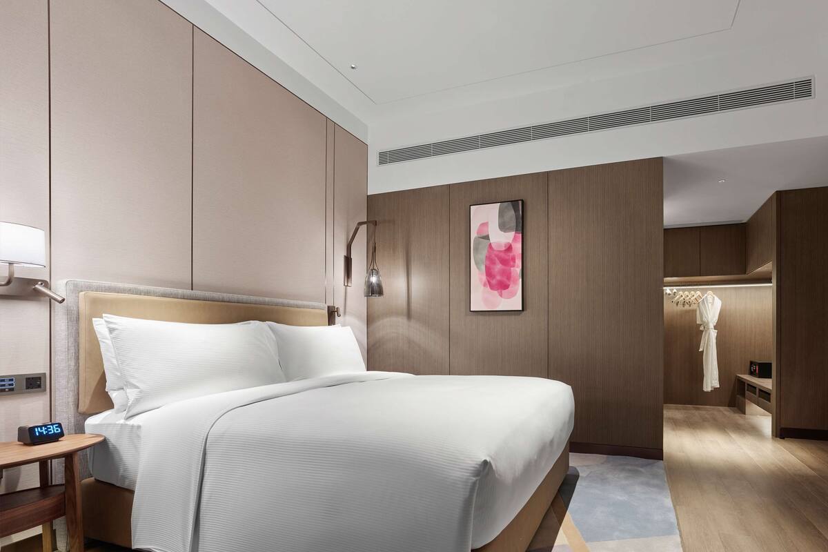 Habitació d'hotel moderna amb un llit gran, una taula de nit amb un rellotge, una làmpada penjada a la paret i un armari amb una bata.