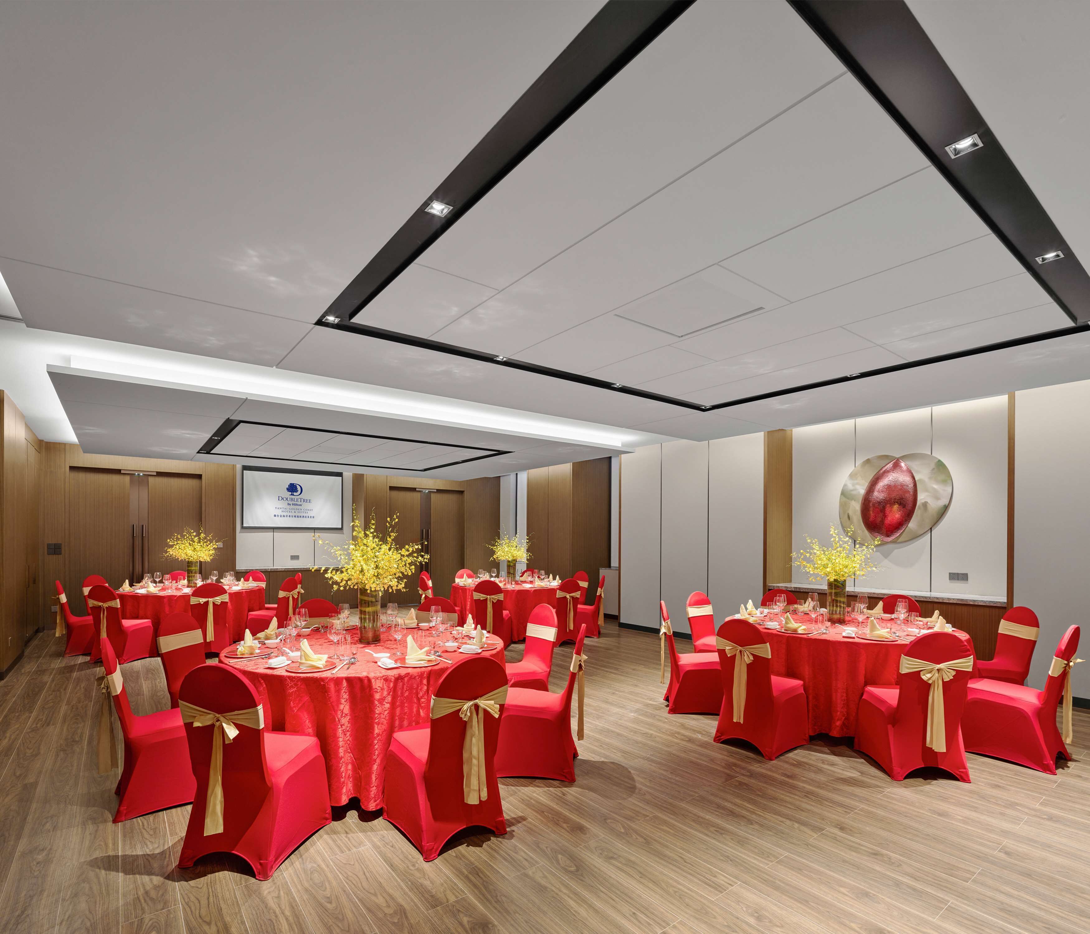 A conference room with multiple tables, red chairs with gold sashes, and a projector screen on the wall.