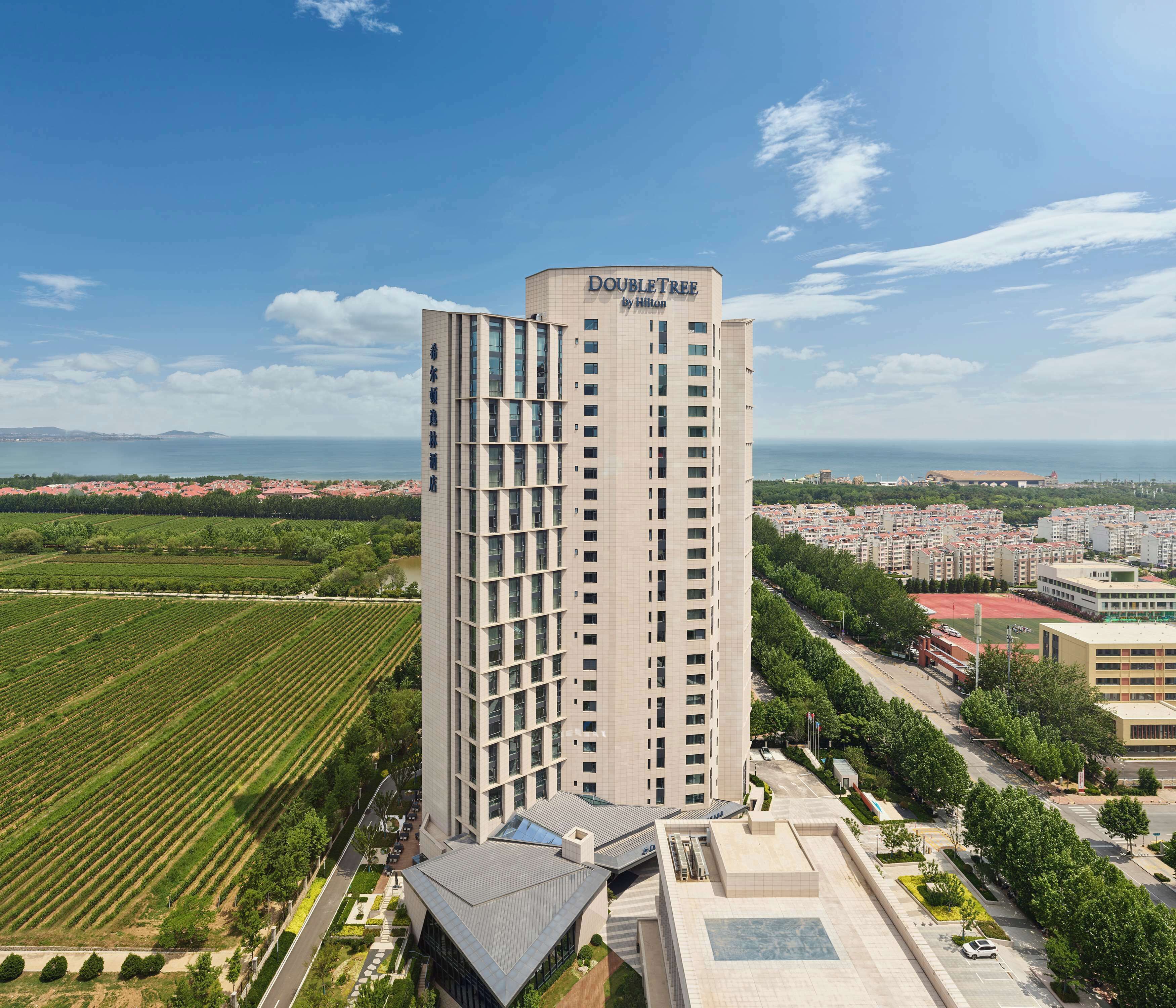 A high-rise hotel named 'DOUBLETREE by Hilton' with a modern architectural design, surrounded by greenery and a view of the sea.