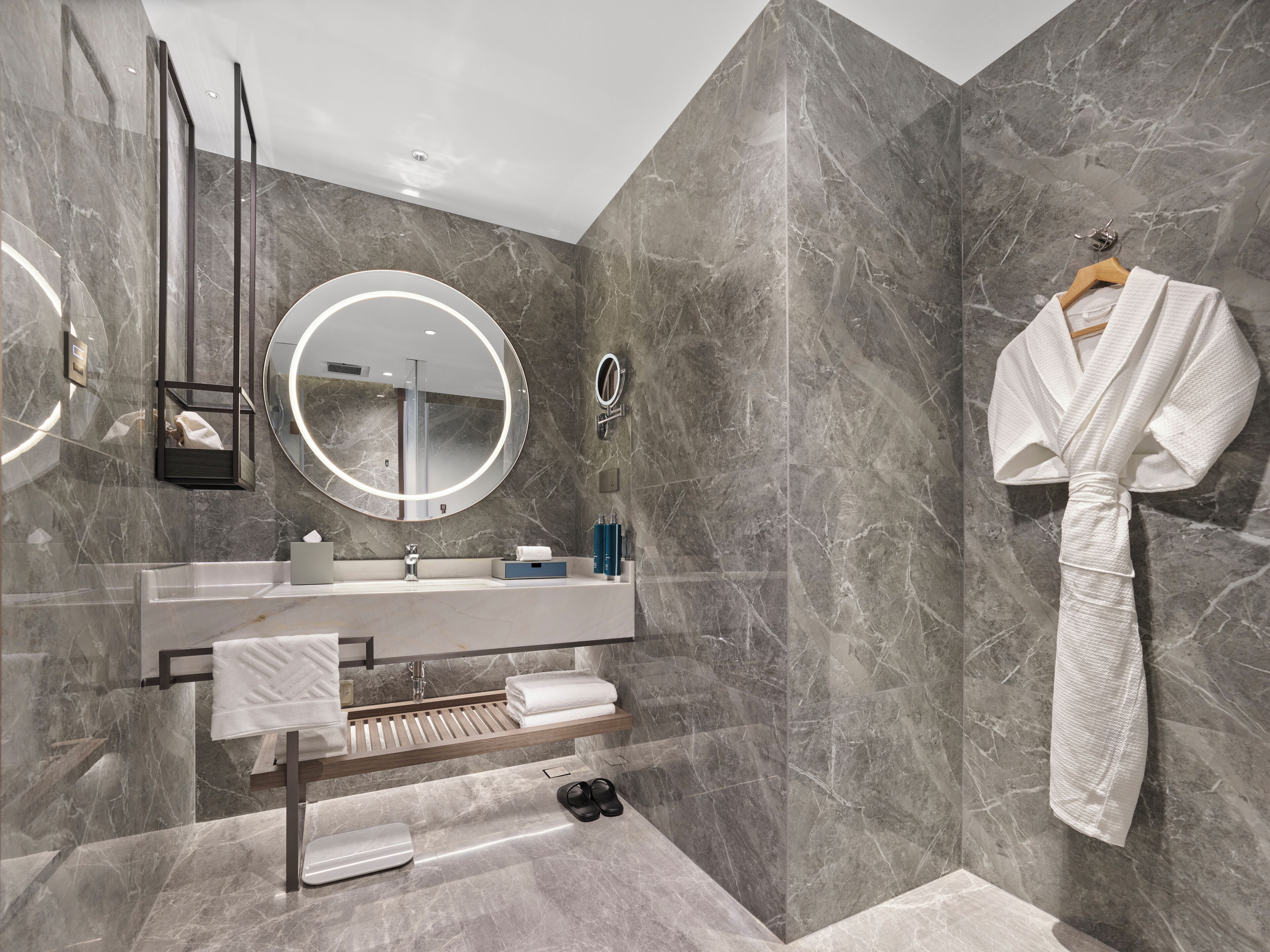 A modern bathroom with a marble wall, a round mirror, a wooden towel rack, and a white robe hanging on the wall.