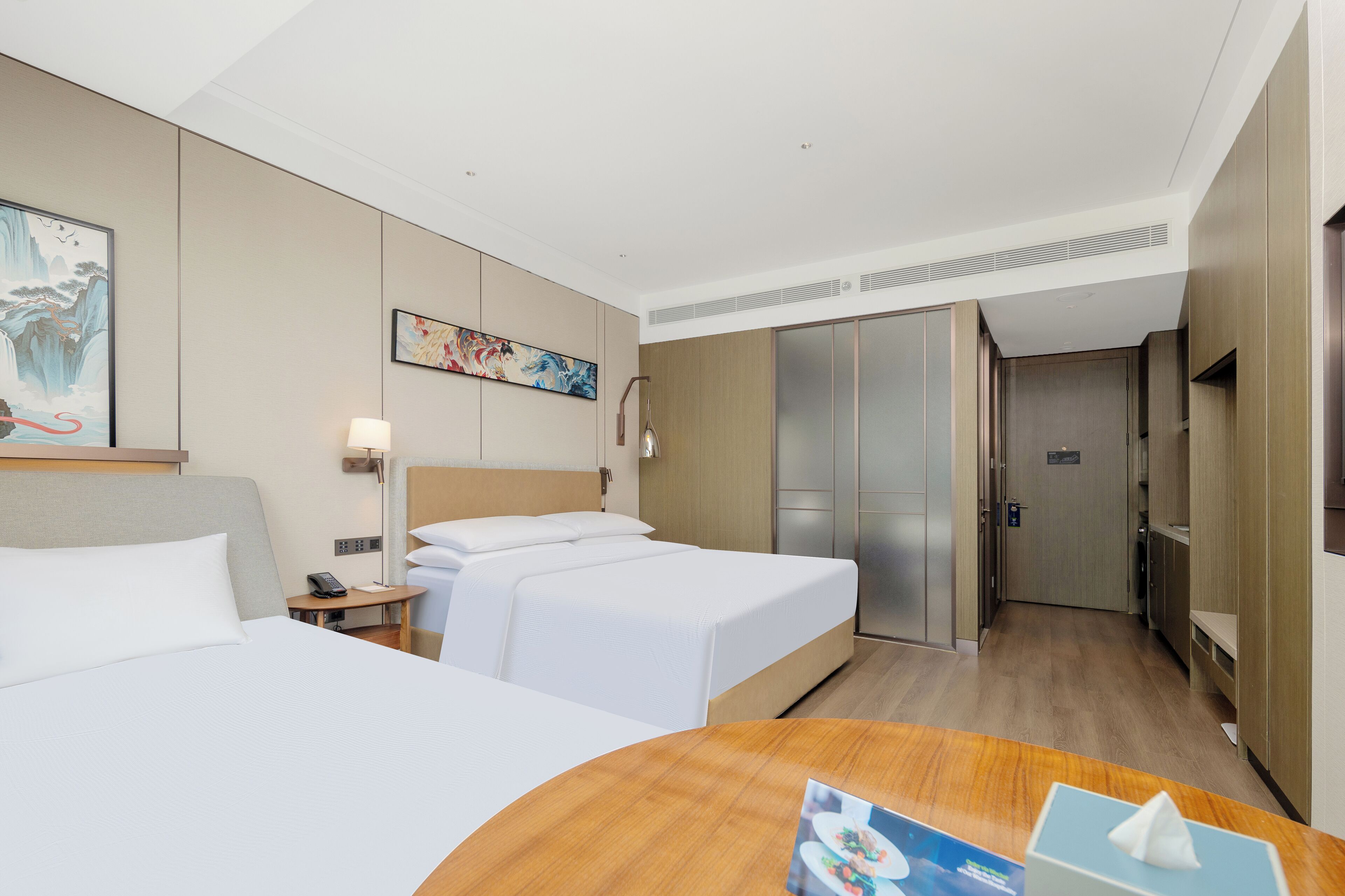 A hotel room with two beds, a wooden desk, a telephone, and artwork on the walls.