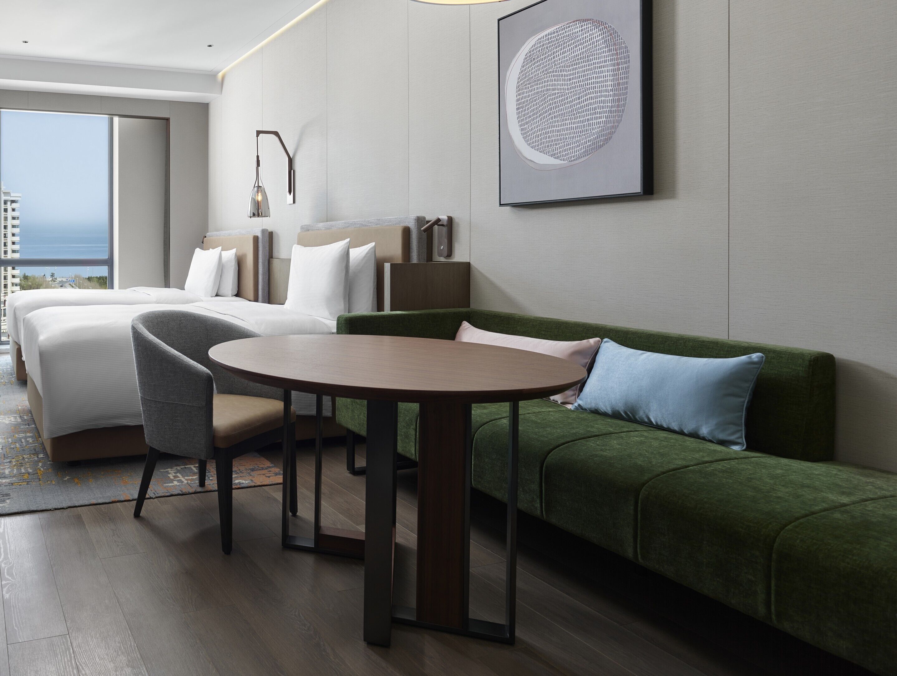A hotel room with a bed, a green sofa, a round table, and a large window offering a view of the city.