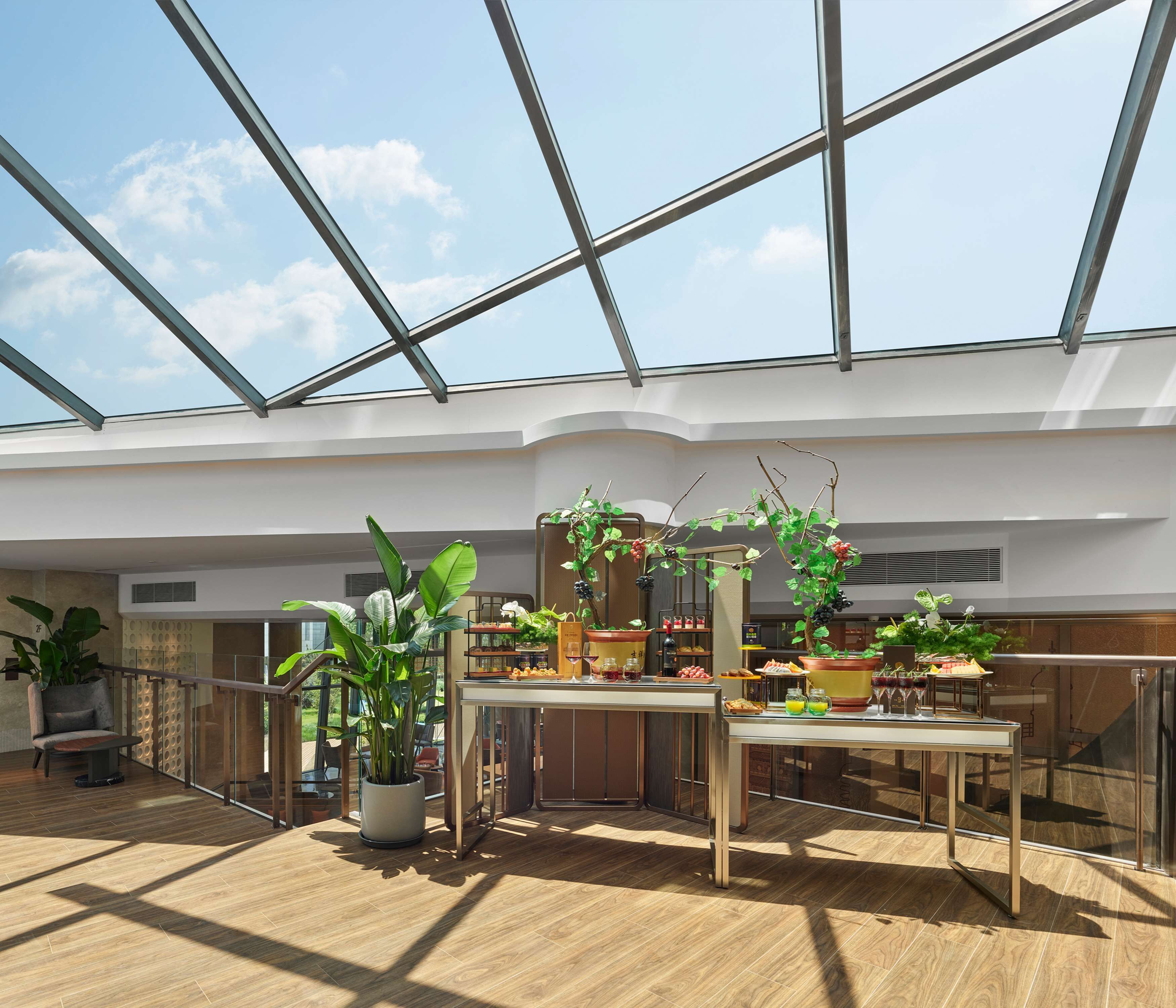 A spacious indoor area with a high ceiling and large windows, featuring a central table with various items and potted plants.