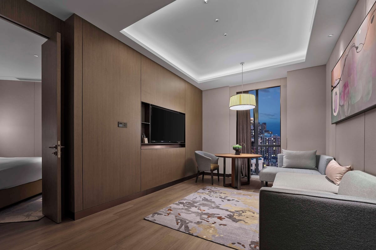 Una habitació d'hotel moderna amb un llit gran, zona de seients amb un sofà i cadires, televisió de pantalla plana penjada a la paret i vista a la ciutat per la finestra.
