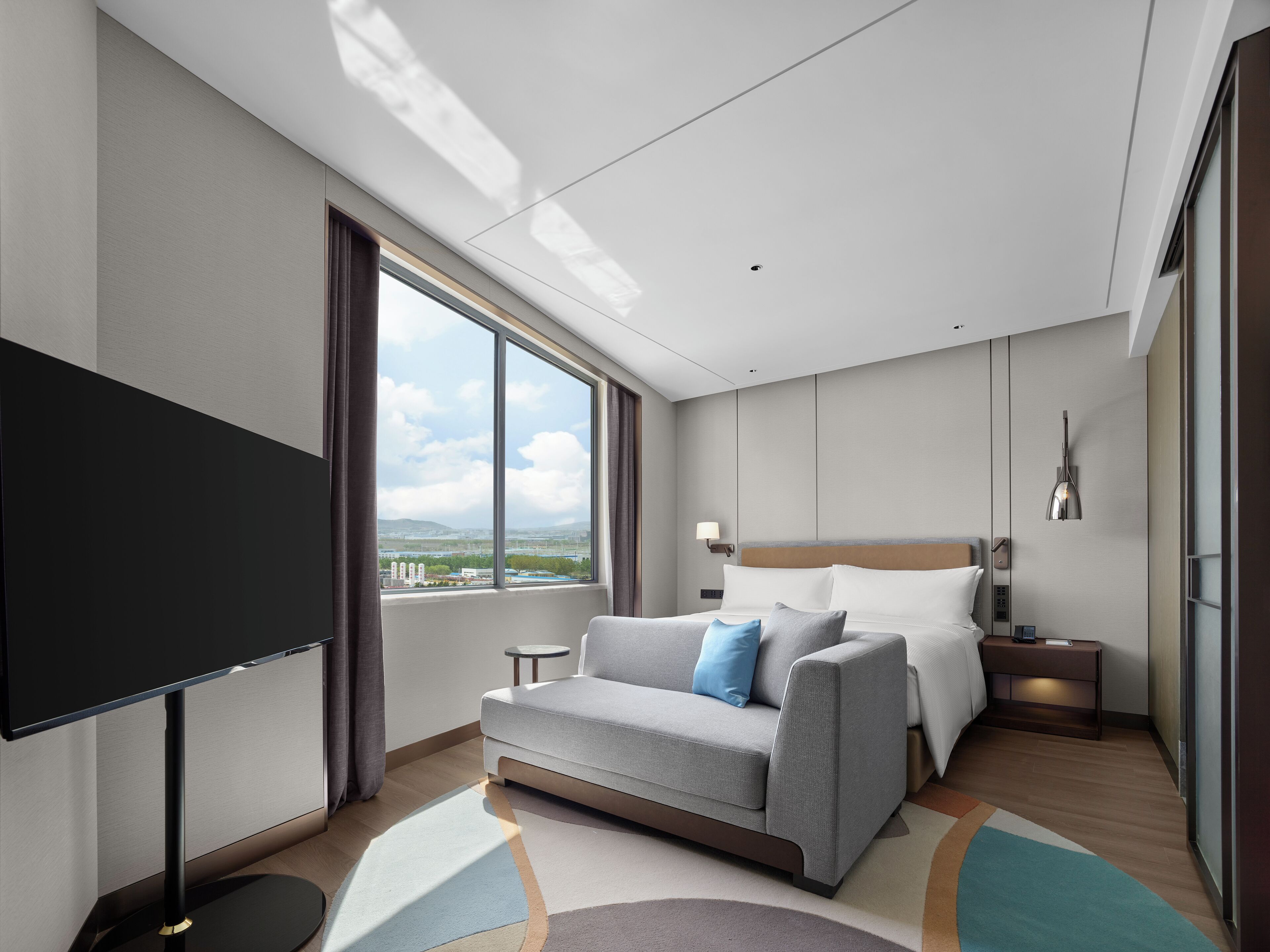 A modern hotel room with a large bed, a sofa, a TV, and a view of the city.