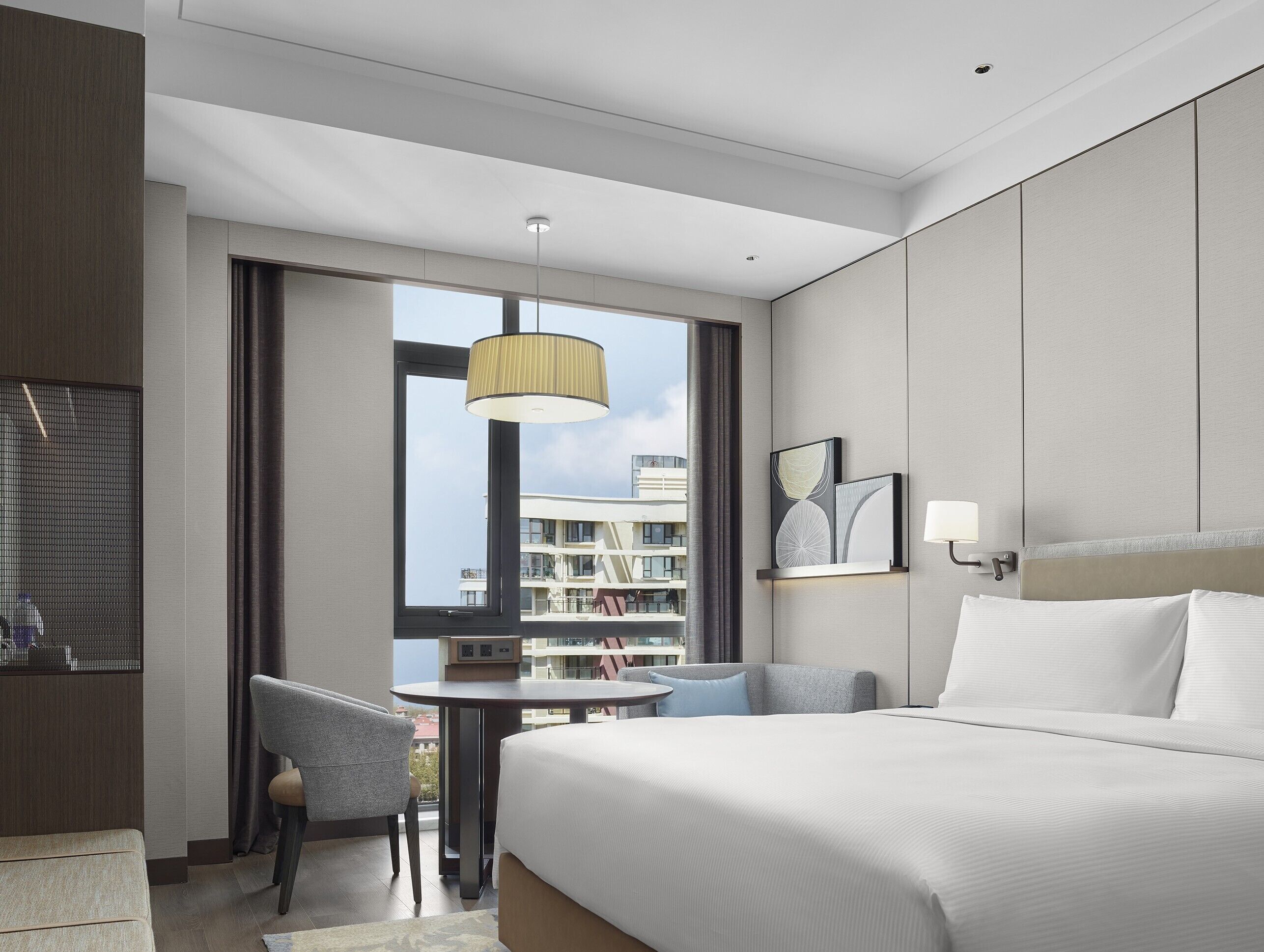 Una habitación de hotel moderna con una cama grande, un escritorio con una silla, una mesa redonda y vistas de la ciudad a través de un ventanal amplio.
