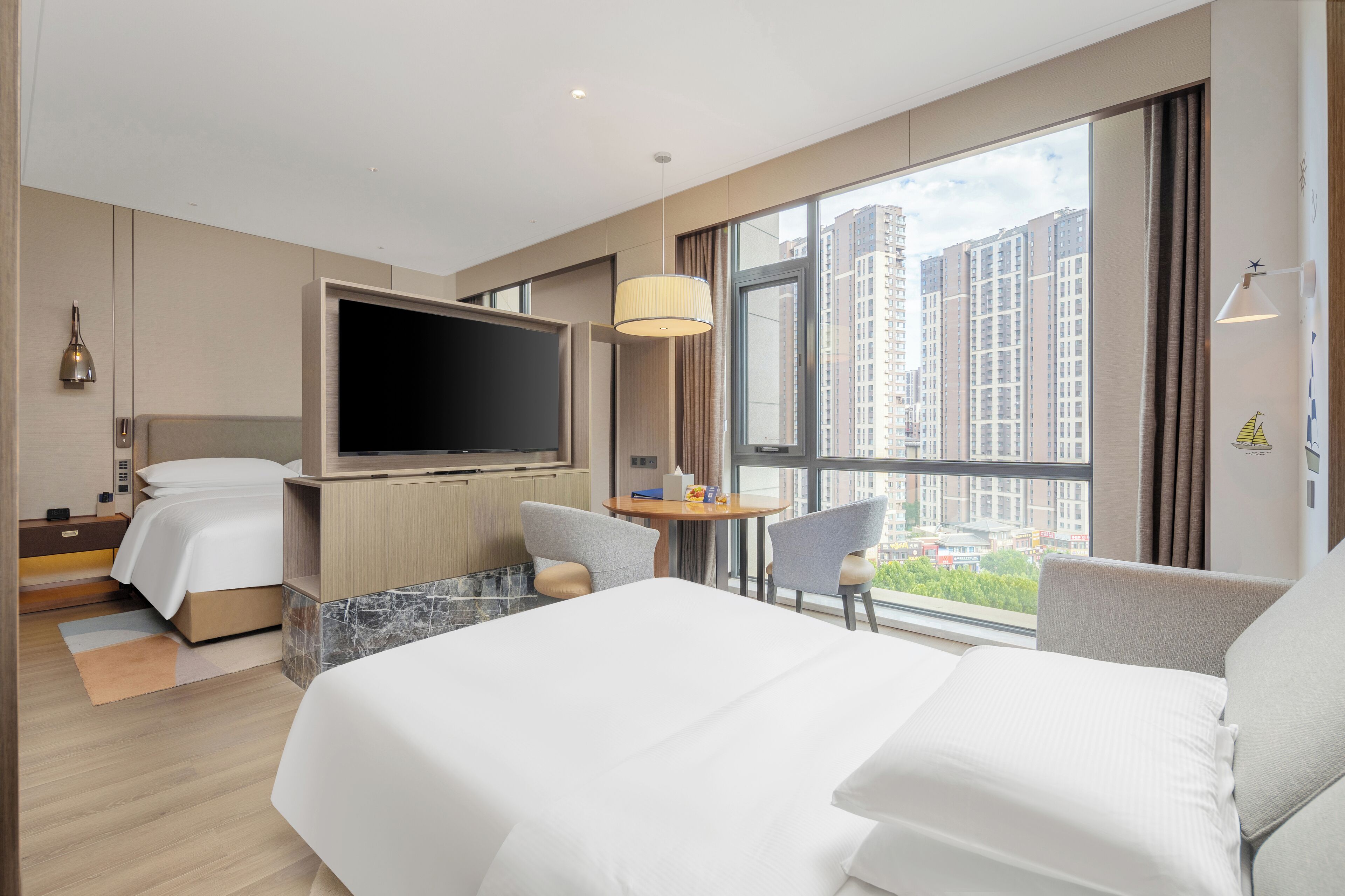 A modern hotel room with a large bed, a flat-screen TV, a dining table, and a view of tall buildings.