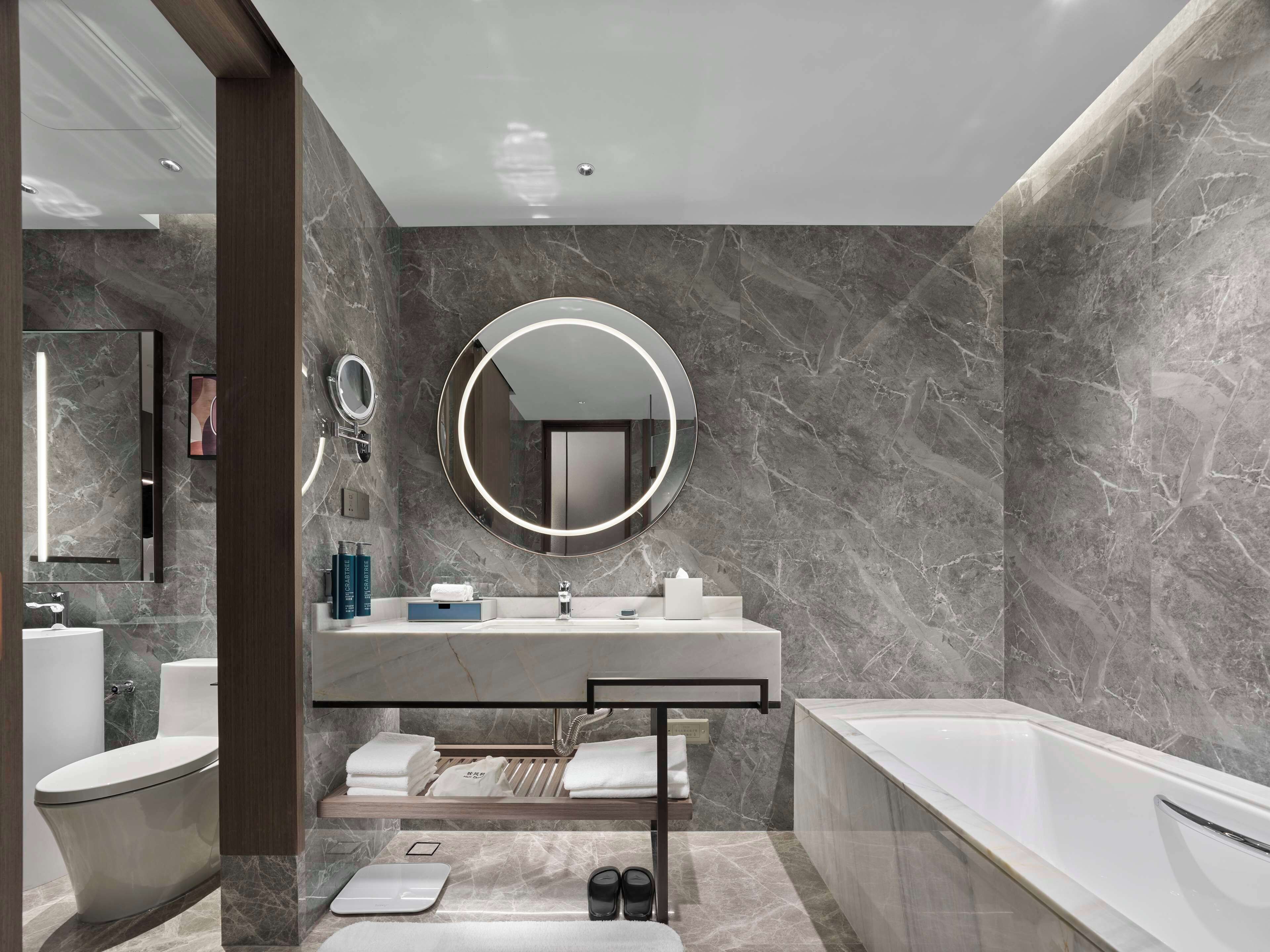 A modern bathroom with a marble wall, a round mirror, a bathtub, and a vanity with a sink and towel rack.