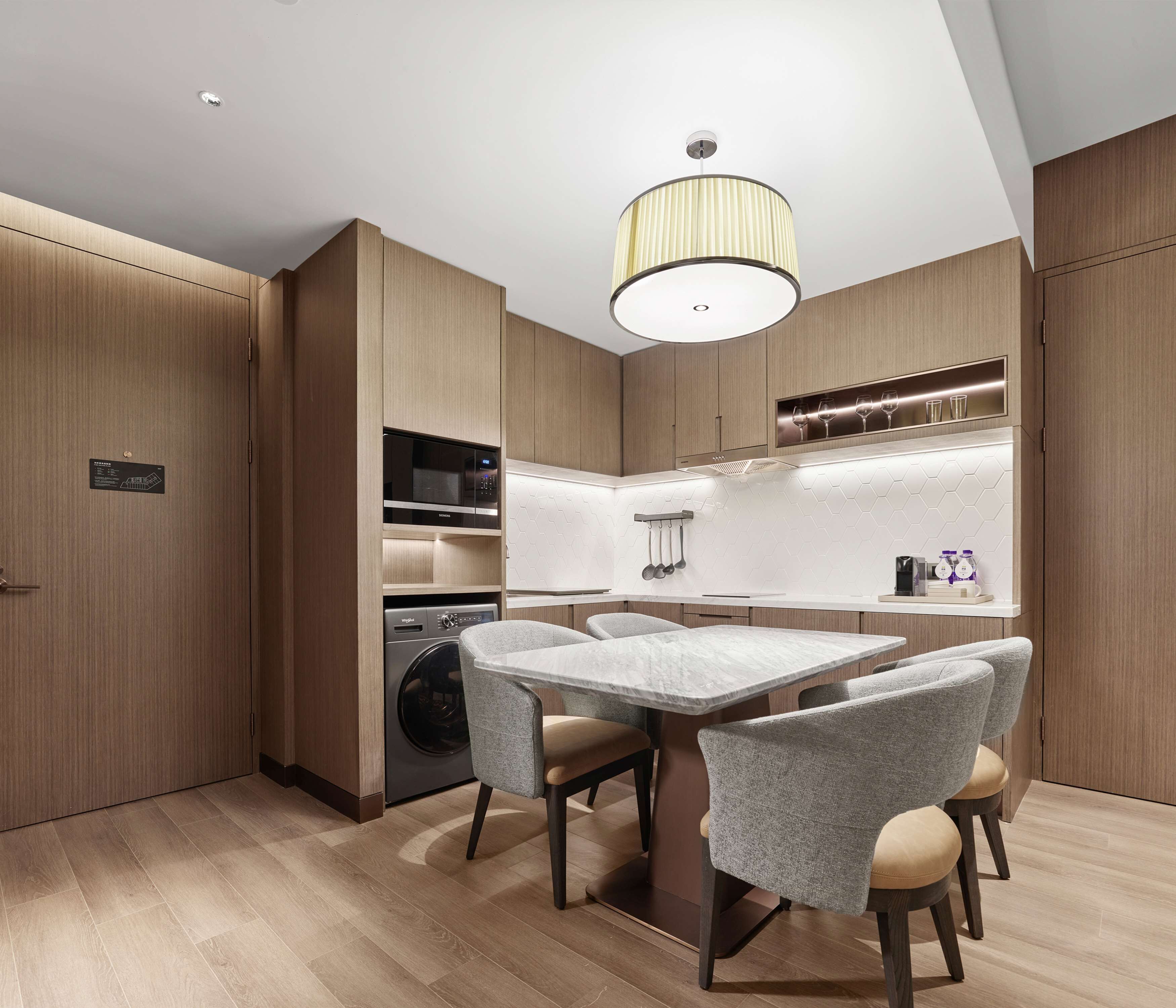 A modern kitchen with wooden cabinetry, a built-in microwave, a coffee machine, and a dining area with a marble table and grey chairs.
