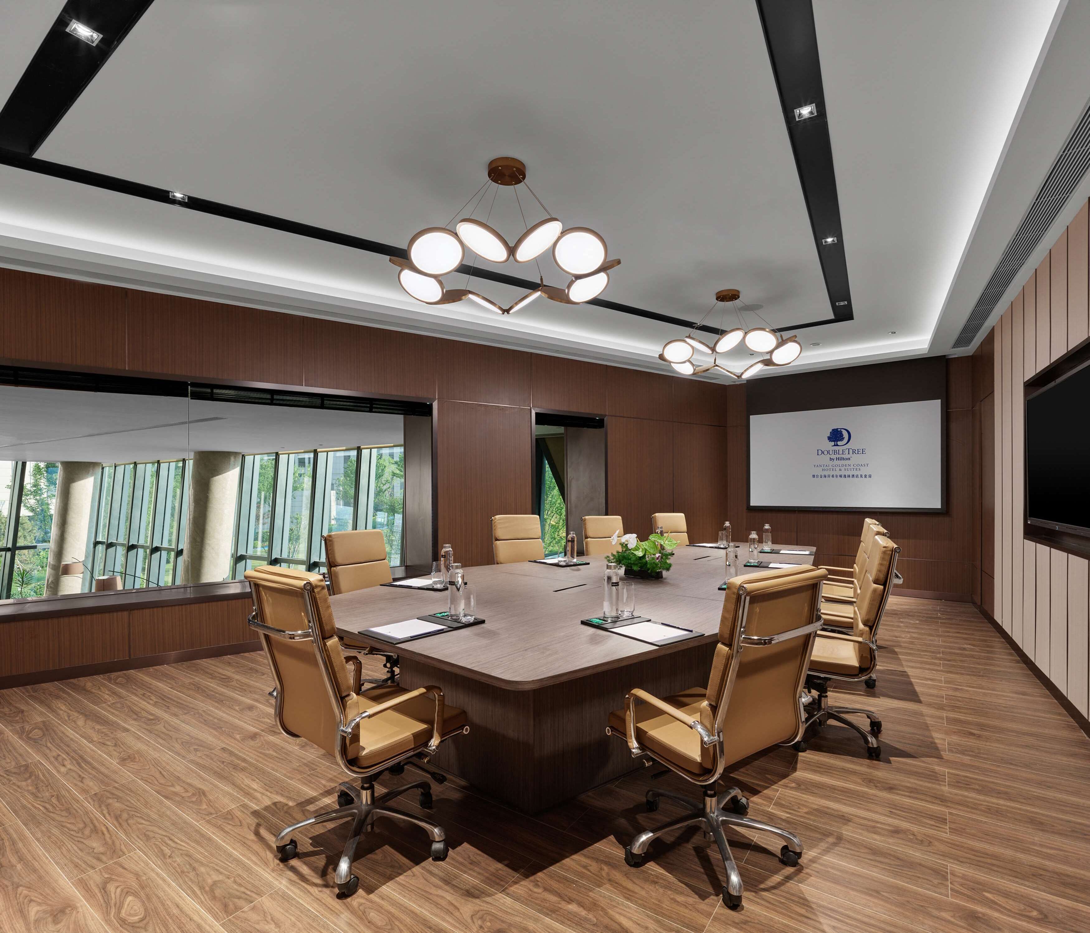 A conference room with a large table, six chairs, a projector screen, and a large window with a view of greenery.