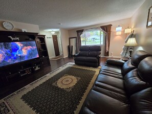 A living room with a flat-screen TV, a leather sofa, a patterned rug, and a window with curtains. - Classy and cozy Ranch Style House, huge fenced backyard and close to everything. (Boise)
