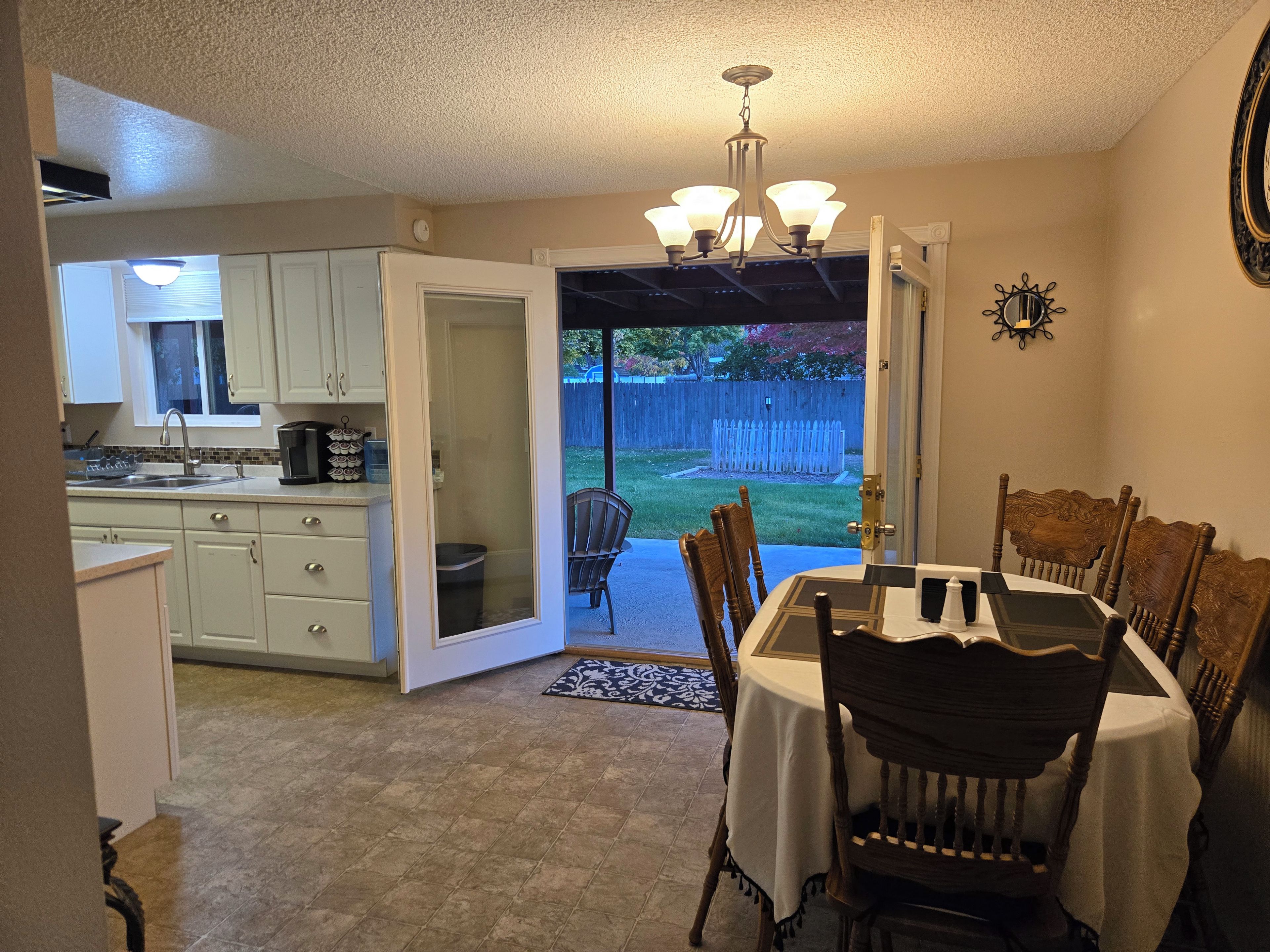 A dining area with a table and chairs, a kitchen in the background, and a view of a backyard with a wooden fence.