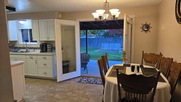 A dining area with a table and chairs, a kitchen in the background, and a view of a backyard with a wooden fence.