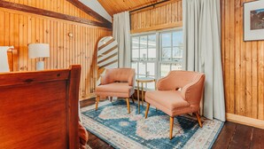 A cozy room with wooden walls, a large window with curtains, two matching armchairs, a small round table, and a patterned rug.