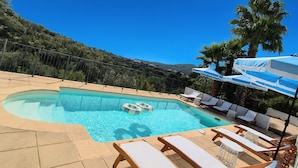 A swimming pool with lounge chairs and umbrellas, surrounded by a hilly landscape.