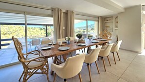 A dining area with a large table set for six, surrounded by wicker chairs, with a view of greenery and hills through large windows.