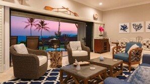 A spacious living area with wicker furniture, a large TV screen displaying a sunset view, and a dining area with a table and chairs. - Luxury Gated Oceanfront Condo on Ground Floor (Kauai County)