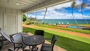 A covered patio with a table and chairs, a pool, and a view of the ocean and palm trees. - Luxury Gated Oceanfront Condo on Ground Floor (Kauai County)