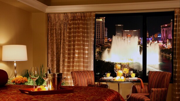 A hotel room with a bed, a table with a tray of items, two chairs, a lamp, and a view of a cityscape with a fountain.