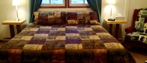 A bed with a quilt featuring animal prints, flanked by two bedside tables with lamps, a chair with a patterned cushion, and a window with curtains.