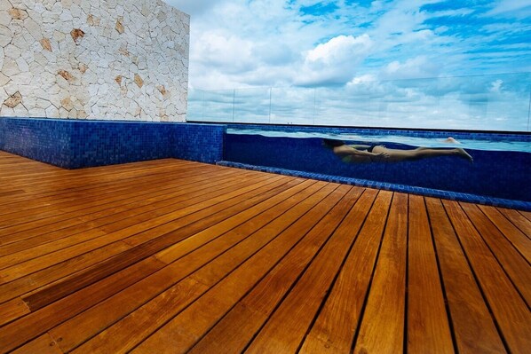 A wooden deck with a pool, a stone wall, and a clear sky.