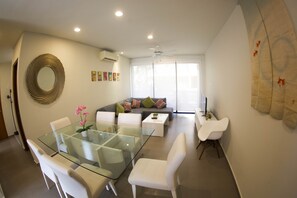 Uma sala de estar moderna com uma mesa de jantar de vidro, cadeiras brancas, um sofá cinza com almofadas coloridas e um espelho grande na parede.