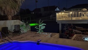 Een zwembad bij nacht met blauwe verlichting, omringd door palmbomen en een houten terras met stoelen.