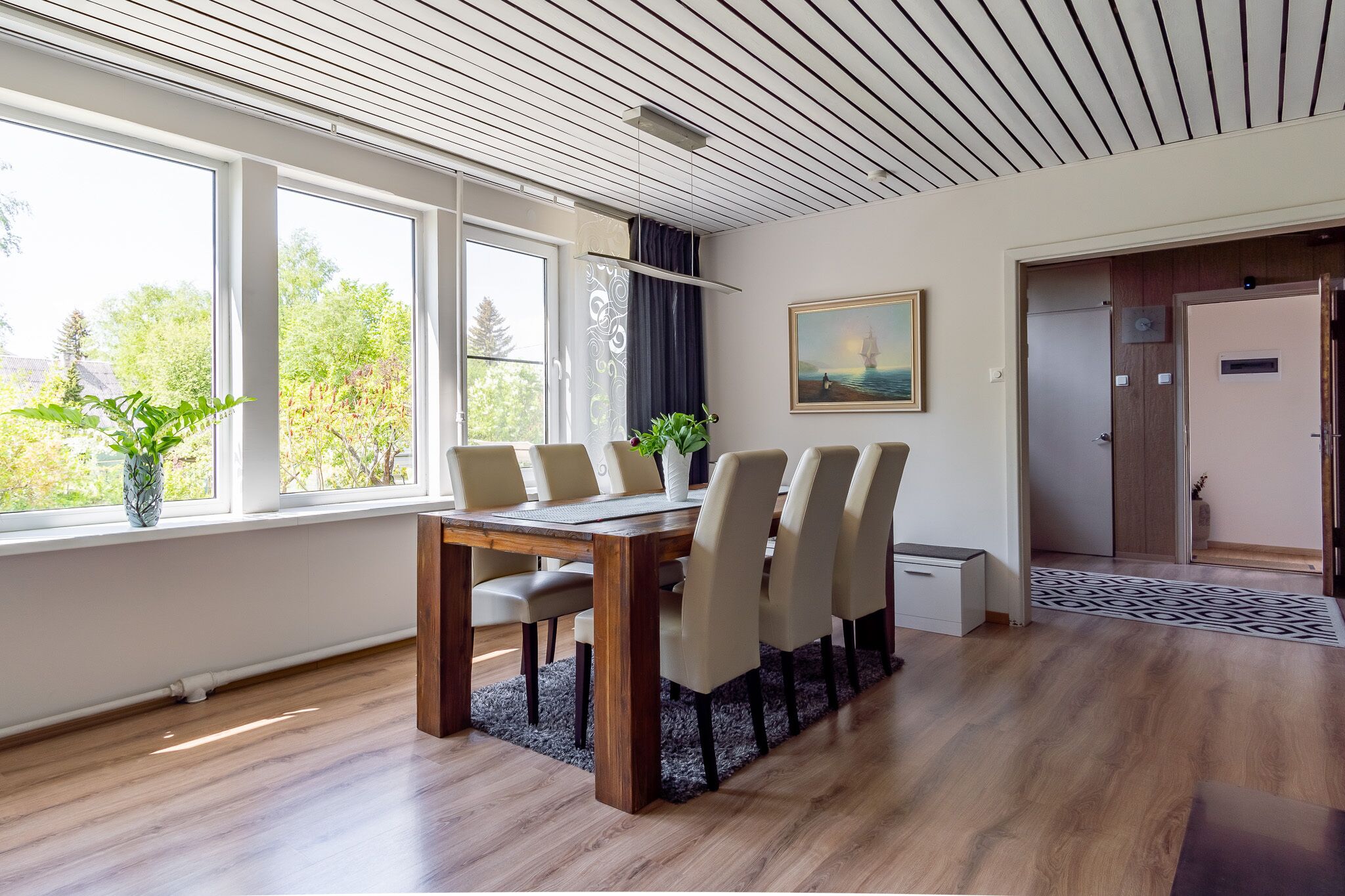 A modern dining area with a wooden table and chairs, large windows, and a painting on the wall.