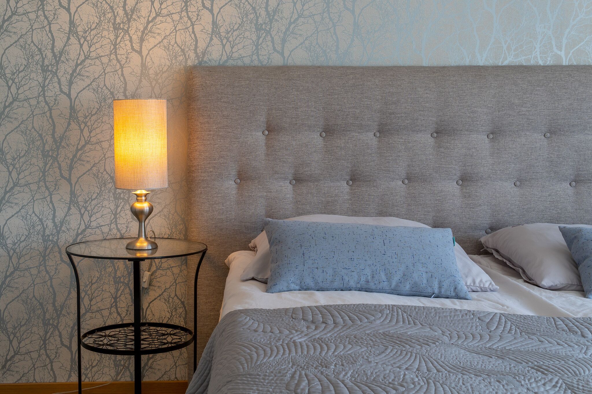 Un lit bien fait, doté d’une tête de lit rembourrée de couleur grise, une table de chevet ronde avec une lampe, et un mur orné d’un motif.