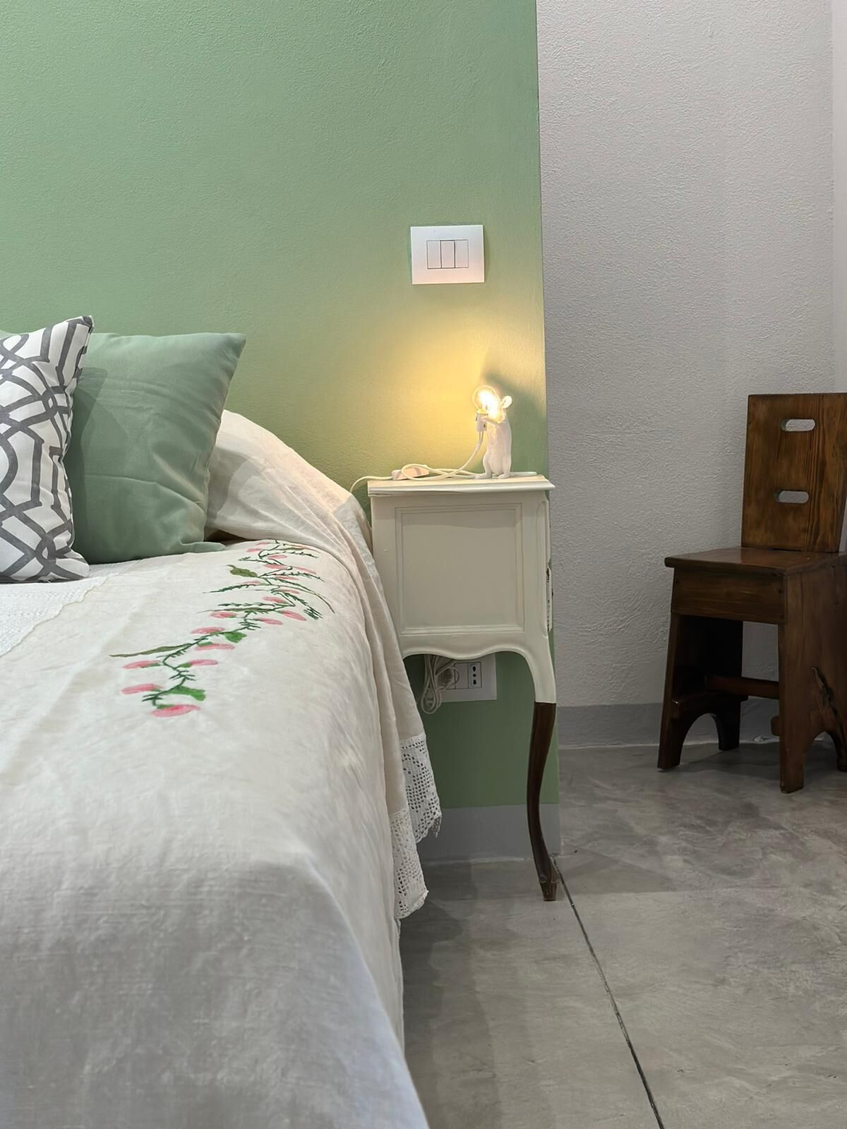 Une chambre à coucher avec un lit, une table de chevet avec une lampe, une chaise en bois et un interrupteur mural.