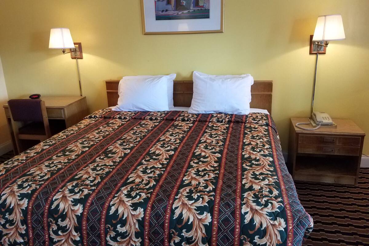 A hotel room with a bed, two bedside lamps, a desk, a chair, a phone on the nightstand, and a framed picture on the wall.
